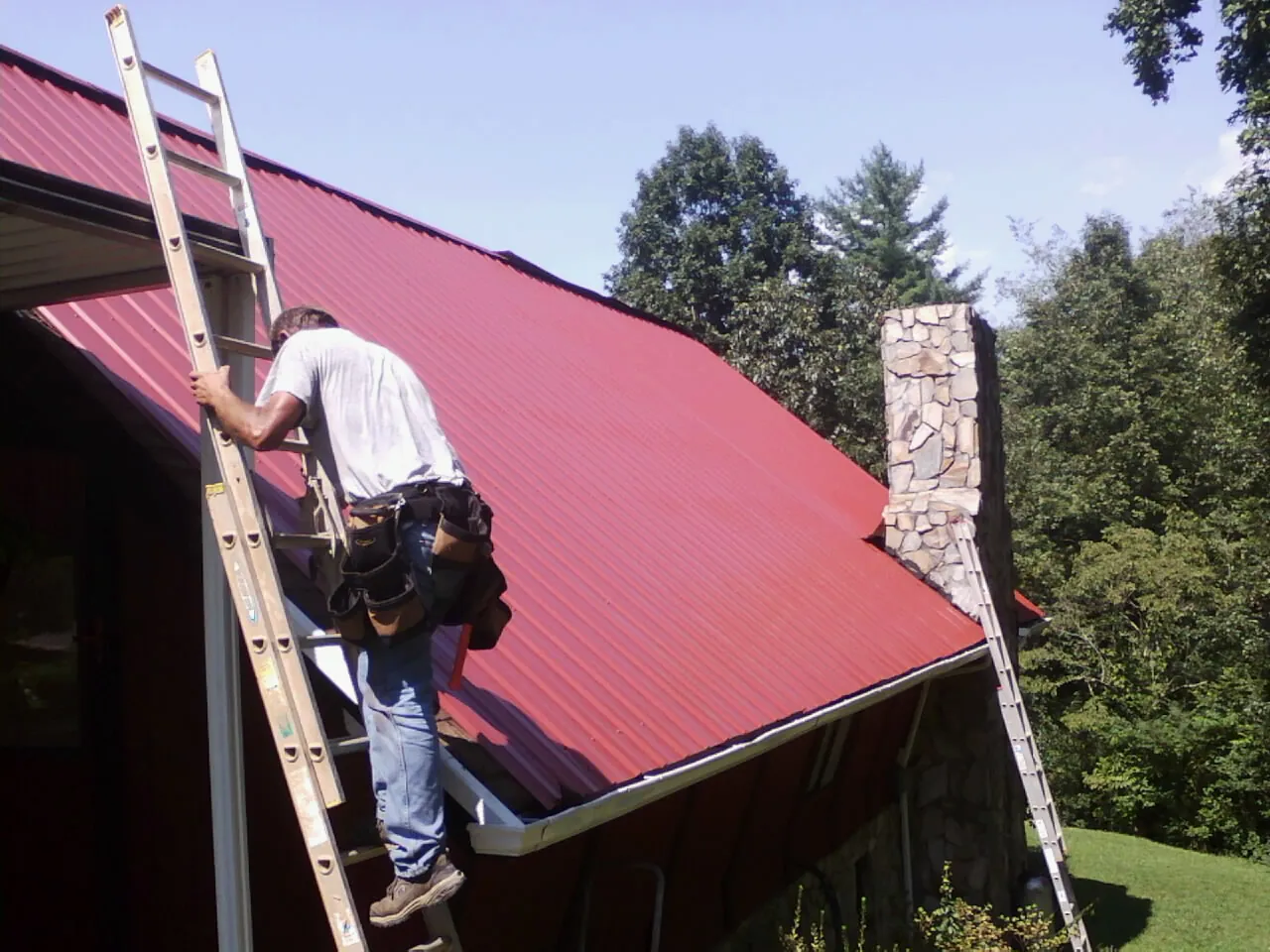 Roofer on a ladder beside a red metal roof and stone chimney — Soffit & Fascia Repair by Roofing Brothers in Bridgton