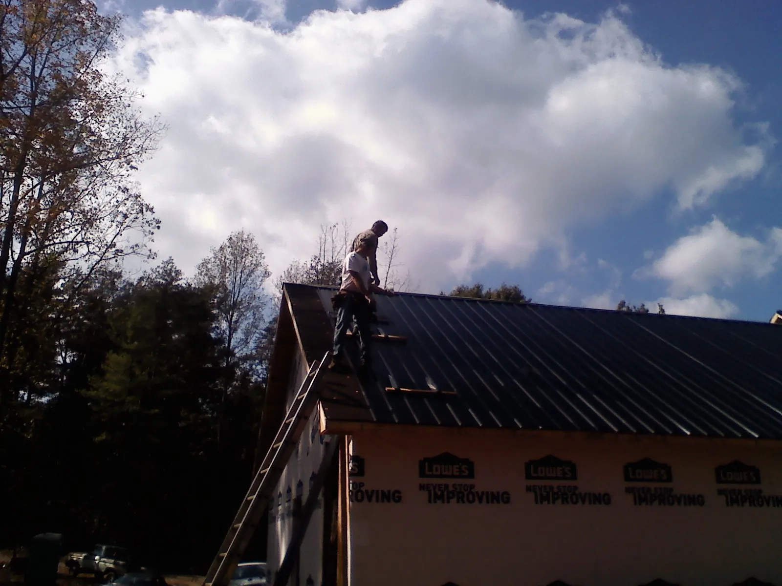 Roofer fastening dark metal panels high against a cloudy sky, from a Roof Inspection job in Bridgton